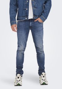Uomo che indossa una giacca di denim blu, una camicia bianca, jeans slim-fit blu scoloriti e sneakers bianche con dettagli neri e verdi, in piedi con le mani nelle tasche.