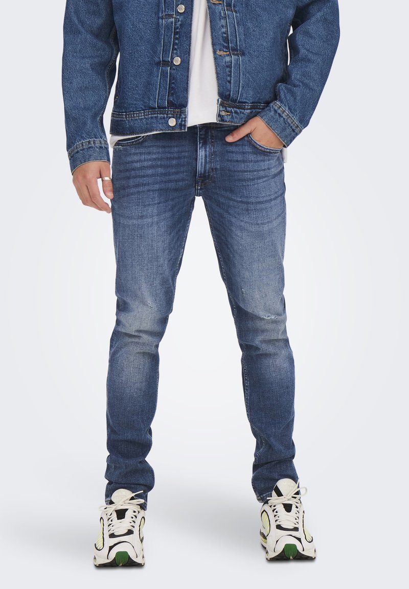 Uomo che indossa una giacca di denim blu, una camicia bianca, jeans slim-fit blu scoloriti e sneakers bianche con dettagli neri e verdi, in piedi con le mani nelle tasche.