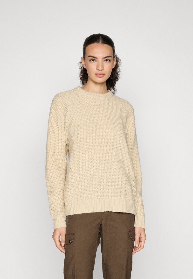 Beige gebreide trui met textuur, voorzien van een geribde halslijn en boorden, met een ontspannen pasvorm en raglanmouwen. Gecombineerd met bruine cargobroek.