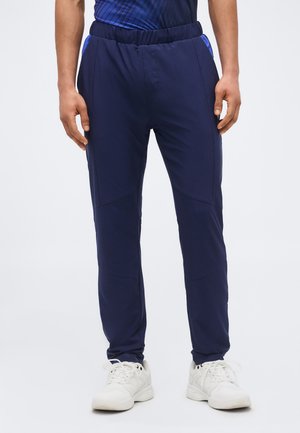 Bărbat purtând pantaloni sportiv bleumarin și adidași albi, stând cu mâinile relaxate pe lângă corp pe un fundal simplu.