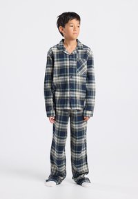 Niño de pie, vistiendo pijama de manga larga a cuadros en azul y blanco con pantalones a juego y calcetines blancos, mirando de lado contra un fondo blanco.