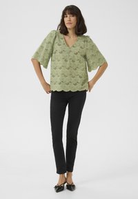 Blusa verde chiaro con scollatura a V, maniche a sbuffo e orlo ondulato, decorata con un motivo floreale a taglio. Abbinata a pantaloni neri elasticizzati.