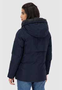 Donna con capelli scuri che indossa una giacca con cappuccio blu marina e jeans blu, mostrata di spalle su uno sfondo neutro.