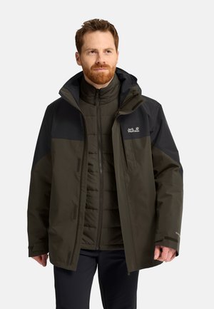 Mann mit rotem Bart, der mehrere dunkelgrüne und schwarze Outdoor-Jacken trägt, steht vor einem einfarbigen Hintergrund.