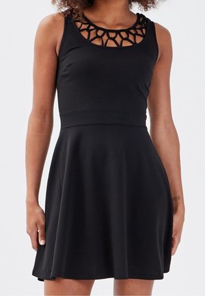 Femme portant une robe noire sans manches avec une taille cintrée et un détail d'encolure découpée en cercle sur fond uni.