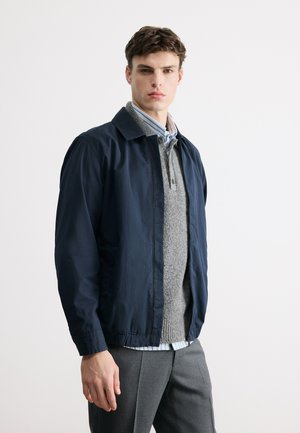 Ung mand iført navyblå jakke over grå sweater og stribet skjorte med krave, stående foran en ensfarvet lys baggrund.