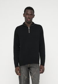 Jaunas vyras, vilkintis juodą megztinį su ketvirčio užtrauktuku ir pilkas džinsinis kelnes, stovi priešais vientisą baltą foną.