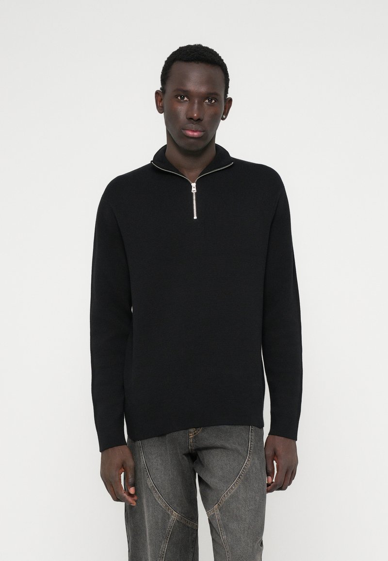 Jaunas vyras, vilkintis juodą megztinį su ketvirčio užtrauktuku ir pilkas džinsinis kelnes, stovi priešais vientisą baltą foną.