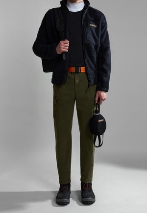 Osoba ubrana w czarną polarkową kurtkę, biały golf, czarną koszulę, oliwkowe spodnie typu cargo, czarne buty, pomarańczowy pasek, niosąca czarny plecak i butelkę z wodą.