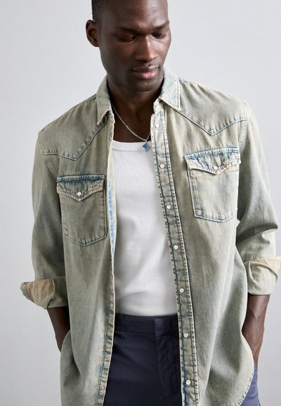 Camisa de denim azul claro con un yugo de estilo western, dos bolsillos en el pecho, botones a presión y mangas arremangadas, llevada sobre una camiseta sin mangas blanca.