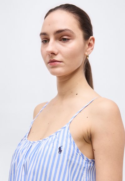 Νεαρή γυναίκα με καστανά μαλλιά σε αλογοουρά φοράει μπλε και λευκό ριγέ αμάνικο τοπ με ένα μικρό σκοτεινό λογότυπο στο στήθος.