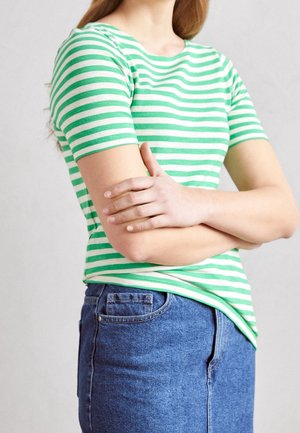 Personne portant une chemise à rayures vertes et blanches à manches courtes et un jean bleu, les bras croisés devant un fond uni.