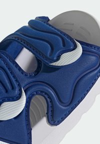 Niebieska gumowa sandałka z teksturowaną górą o zaokrąglonych wzorach, regulowanymi paskami i białymi akcentami. Biała, solidna podeszwa z wzorem zapewniającym przyczepność.