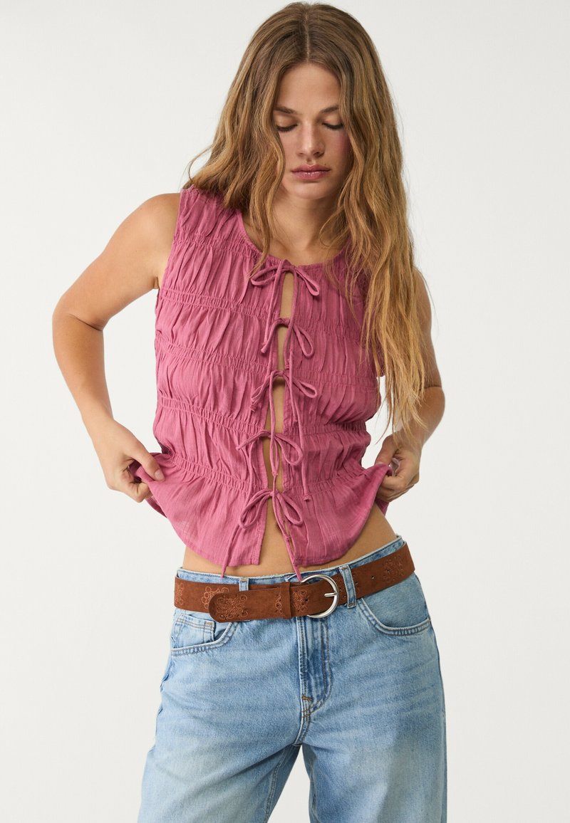 Rosa, krinklat tygtop med en frontknut, ärmlös design och elastisk midja; bärs med blå denimjeans och ett brunt bälte.
