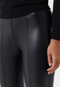 Schwarze Leggings aus glänzendem Kunstleder, mit vertikalen Nähten, glatter Textur und eng anliegendem Design. Hand ruht auf dem Oberschenkel.