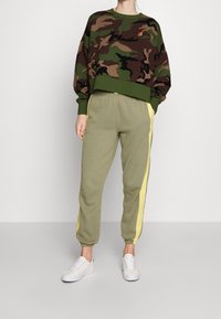 Camo-patterned cropped sweatshirt in shades of green and brown, paired with olive joggers featuring yellow side stripes and white sneakers.