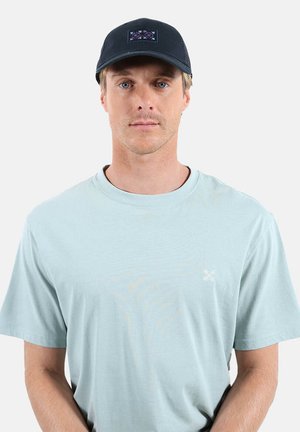 Homme portant un t-shirt bleu clair à encolure ronde et une casquette noire avec un motif brodé, debout devant un fond uni et clair.