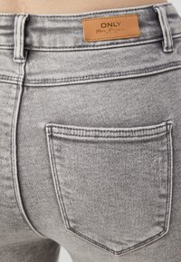 Jeans de mezclilla gris con una cinturilla alta, bolsillos traseros y una etiqueta de cuero marrón en la parte trasera central. Costuras visibles y detalles de textura.