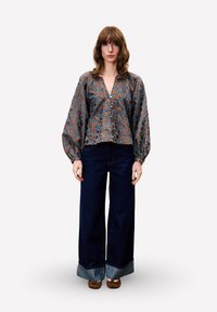 Frau in einer braun-blauen gemusterten Bluse mit voluminösen Ärmeln, weit geschnittenen, dunklen Jeans mit Umschlägen und braunen Schuhen, steht.