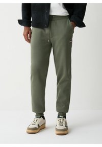 Pantaloni de jogging verde măsliniu dintr-un material moale, cu talie ajustabilă prin șnur, croi conic și o mică etichetă cu logo pe coapsă. Purtați cu adidași albi.