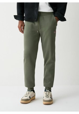 Olivgrüne Jogginghose aus weichem Stoff, mit einem Kordelzugbund, schmalem Schnitt und einem kleinen Logopatch am Oberschenkel. Getragen mit weißen Sneakers.