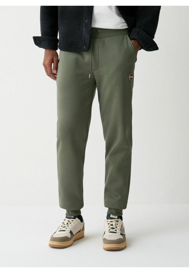 Pantaloni de jogging verde măsliniu dintr-un material moale, cu talie ajustabilă prin șnur, croi conic și o mică etichetă cu logo pe coapsă. Purtați cu adidași albi.