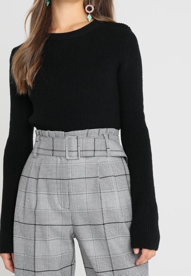 Femme portant un pull à manches longues côtelé noir et un pantalon taille haute gris à carreaux avec une ceinture assortie.