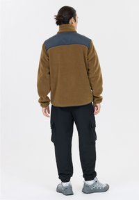 Brun fleecjacka med en mörkblå axelpanel, med långa ärmar och ståkrage, tillsammans med svarta cargobyxor och grå sneakers.