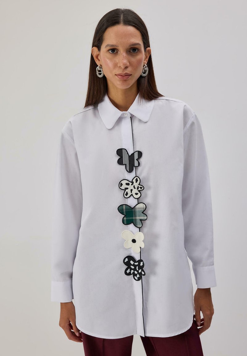 Kobieta ubrana w białą koszulę na guziki z pięcioma wzorzystymi łatkami motyli z przodu, do tego bordowe spodnie i srebrne kolczyki koła.