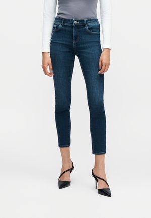 Femme portant un jean slim bleu foncé aux chevilles, un haut gris, des manches longues blanches et des chaussures à talons noirs à bout pointu, debout sur un fond uni.