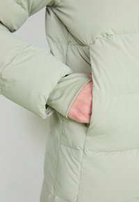 Giacca verde imbottita con una texture morbida, tasche laterali e polsini elasticizzati che si estendono oltre la mano per calore e comfort.
