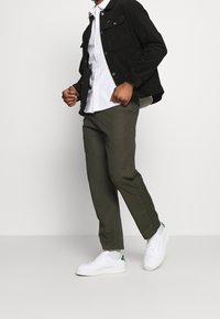 Zwarte corduroy jas, witte blouse, olijfgroene broek en witte sneakers met groene accenten. Verhoogde kraag en details aan de zijnaad.