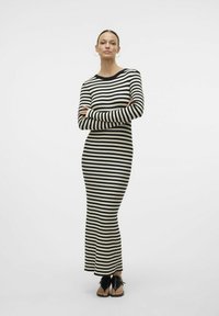 Robe en tricot rayée à manches longues, avec des rayures horizontales noires et blanches, coupe ajustée et texture côtelée. Portée avec des sandales noires.