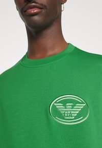 T-shirt in cotone verde con scollatura rotonda e logo ricamato con ali in bianco e verde al centro. Texture morbida.