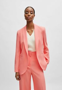 Peach-colored tailored blazer with a shawl collar, paired with matching trousers. Smooth fabric, minimalistic design, featuring side pockets.