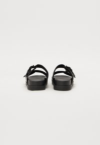 Sorte sandaler med flad sål, som har en bred rem og en spænde detalje. Lavet af syntetisk materiale med en glat tekstur.