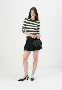 Vrouw in een groen en wit gestreepte trui, zwarte geplooide rok, beige sokken met zwarte hakken, en een kleine zwarte handtas.