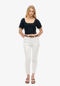 Marineblå kort top med pufærmer, struktureret stof, parret med hvide tætsiddende jeans og et fletbælte i brunt. Brune sandaler fuldender looket.
