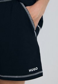 Juodi šortai pagaminti iš minkštos medžiagos, su balta siuvimo linija, turi šonines kišenes ir spausdintą "HUGO" logotipą šalia apatinio krašto.