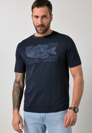 Man met getatoeëerde arm, die een marineblauw T-shirt draagt met een visafbeelding en tekst over offshore vissen, lichte spijkerbroek en een polshorloge, staand tegen een effen achtergrond.