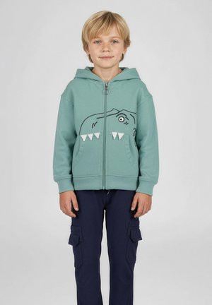 Blond chłopiec w turkusowej bluzie na zamek z motywem twarzy dinozaura oraz granatowych spodniach bojówkach, stojący na jednolitym tle.