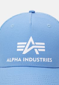 Kék baseball sapka fehér Alpha Industries logóval, amelyen stilizált "A" és három vízszintes csík található a márkaneve felett.
