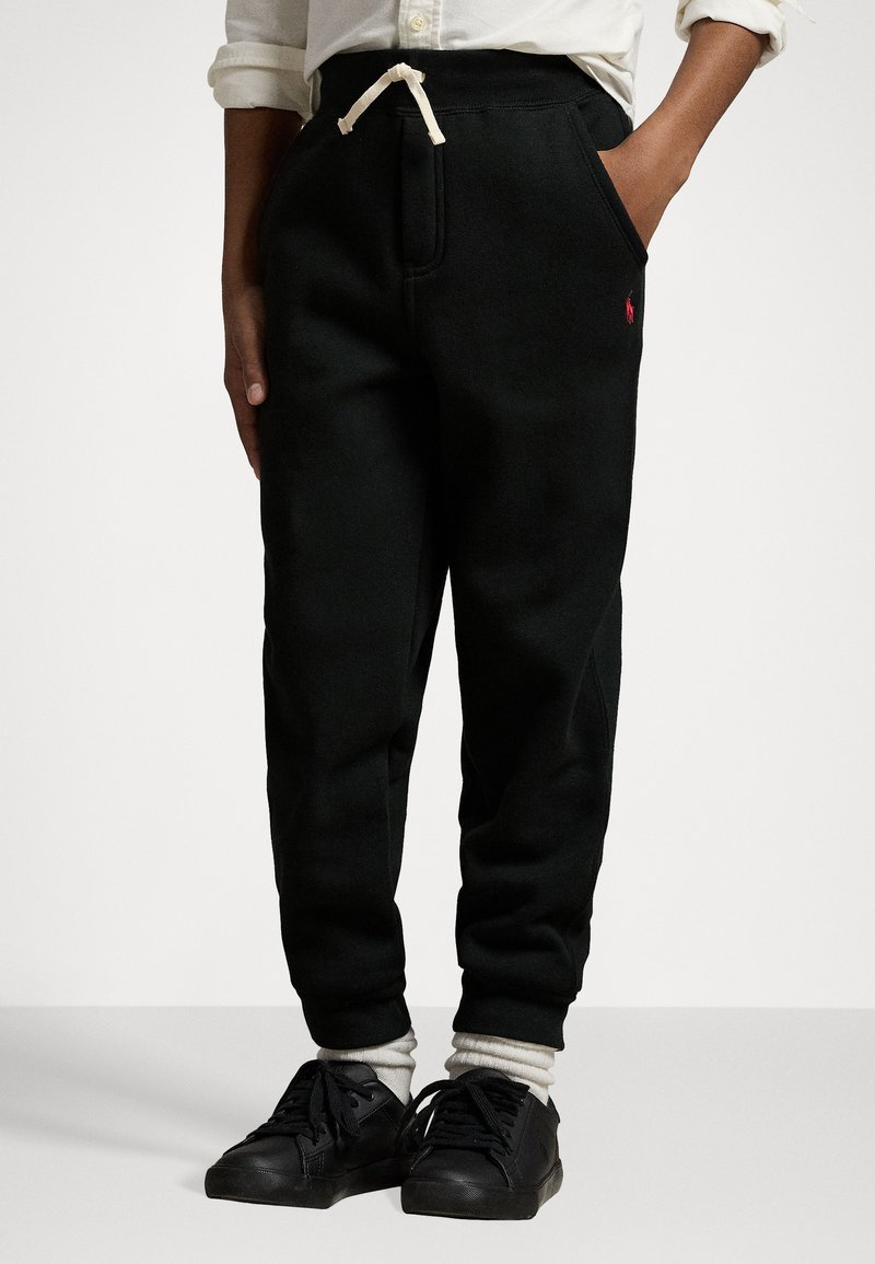Pantalons de sport noirs en tissu doux, dotés d'une taille avec cordon de serrage, de poches latérales et d'un petit logo rouge. Portés avec des baskets noires et des chaussettes blanches.