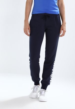 Persona vestida con pantalones deportivos azul marino con texto blanco en la pierna izquierda, zapatillas blancas y una camiseta azul, de pie sobre un fondo blanco.