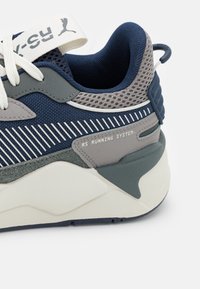 Modré a šedé běžecké boty s síťovinovým svrškem, gumovou podrážkou, texturovanými detaily a brandingem RS Running System na boku. Obsahují ploché bílé tkaničky.