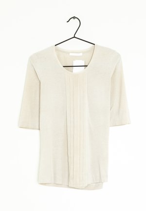 Blouse crème à manches courtes avec plis verticaux sur le devant, suspendue à un cintre en fil de fer noir sur fond blanc.