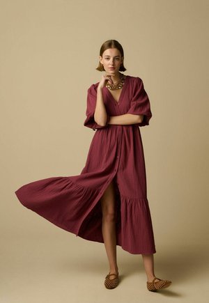 Jeune femme en robe bordeaux fluide et chaussures marron cloutées pose devant un fond beige, la main reposant sur le menton et les jambes visibles à travers la fente.