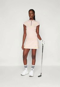 Sieviete, tērpusies gaiši rozā golfa apģērbā un baltā golfa cimdā, tur golfa nūju, stāvot uz vienkārša gaiša fona.