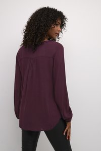 Langärmlige, lila Bluse mit lockerer Passform, weichem Material, plissiertem Rückendetail und abgerundetem Saum, kombiniert mit schwarzen, engen Hosen.
