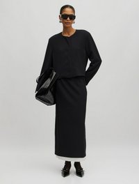 Zwarte korte jas met ronde halslijn, gecombineerd met een lange zwarte rok. Accessoires zijn oversized zwarte zonnebrillen en een glanzende zwarte tote bag.